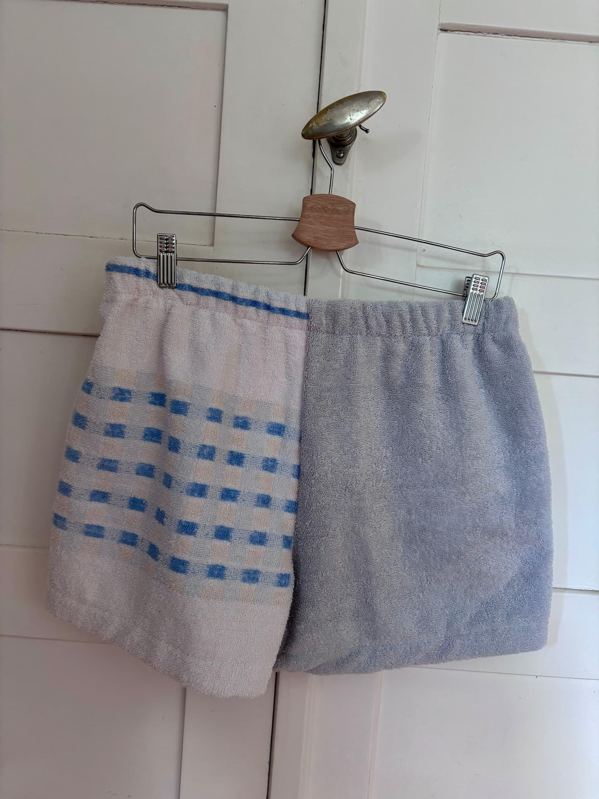 Rückansicht der Frottee-Patchwork-Shorts „Sand & Sun 67“ von Old Wave an einem Kleiderbügel. Diese Seite präsentiert einen weiteren Materialmix aus upgecycelten Handtüchern: Das eine Hosenbein zeigt ein weißes Grunddesign mit quer verlaufenden, hellblauen Streifenmustern, während das andere Hosenbein komplett aus einem schlichten, hellgrau-blauen Frotteestoff gefertigt ist.