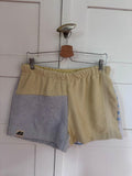 Upcycling-Shorts „Sand & Sun 67“ von Old Wave auf einem Kleiderbügel vor einer weißen Holzwand. Das sommerliche Design aus Frottee-Handtüchern zeigt ein asymmetrisches Patchwork-Muster: Das rechte Hosenbein ist komplett in einem sandfarbenen Beigeton gehalten. Das linke Hosenbein kombiniert ein beiges Element im oberen Bereich mit einem hellgrau-blauen Frotteestoff unten.