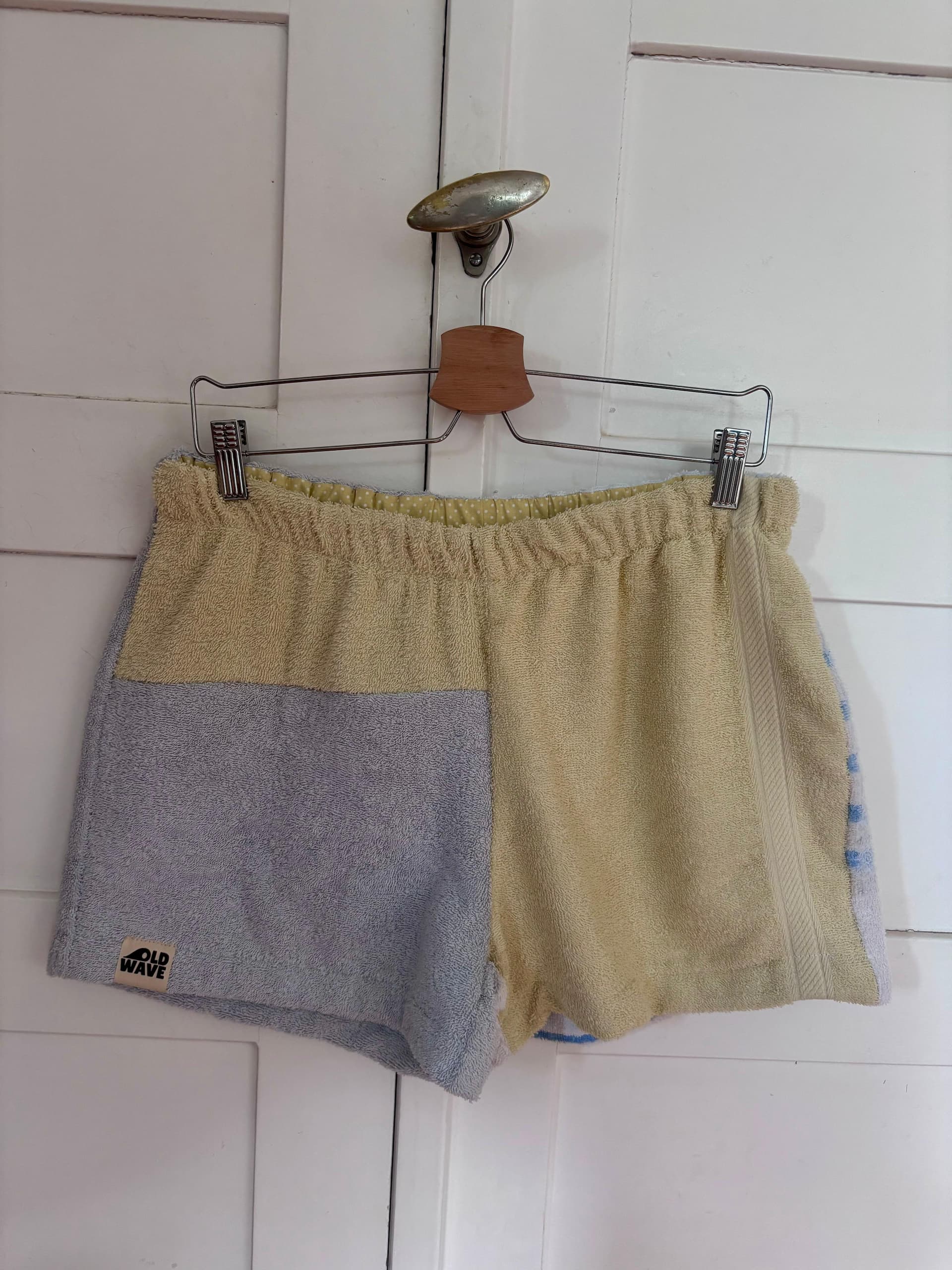 Upcycling-Shorts „Sand & Sun 67“ von Old Wave auf einem Kleiderbügel vor einer weißen Holzwand. Das sommerliche Design aus Frottee-Handtüchern zeigt ein asymmetrisches Patchwork-Muster: Das rechte Hosenbein ist komplett in einem sandfarbenen Beigeton gehalten. Das linke Hosenbein kombiniert ein beiges Element im oberen Bereich mit einem hellgrau-blauen Frotteestoff unten.