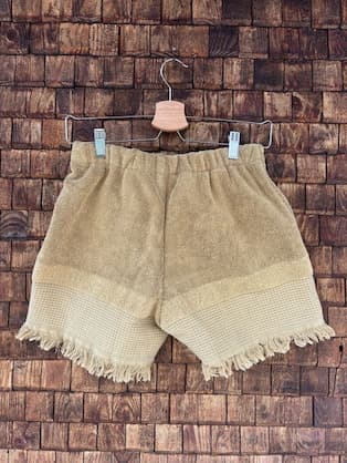 Rückansicht der Upcycling-Frotteeshorts „Sand Fringe ‘91“ von Old Wave auf einem Kleiderbügel vor einer Holzwand. Die Rückseite spiegelt das minimalistische, sandfarbene Konzept wider, bei dem der weiche Frotteestoff im unteren Drittel in eine strukturierte Web-Optik übergeht und mit einem ringsum laufenden Fransensaum abschließt.