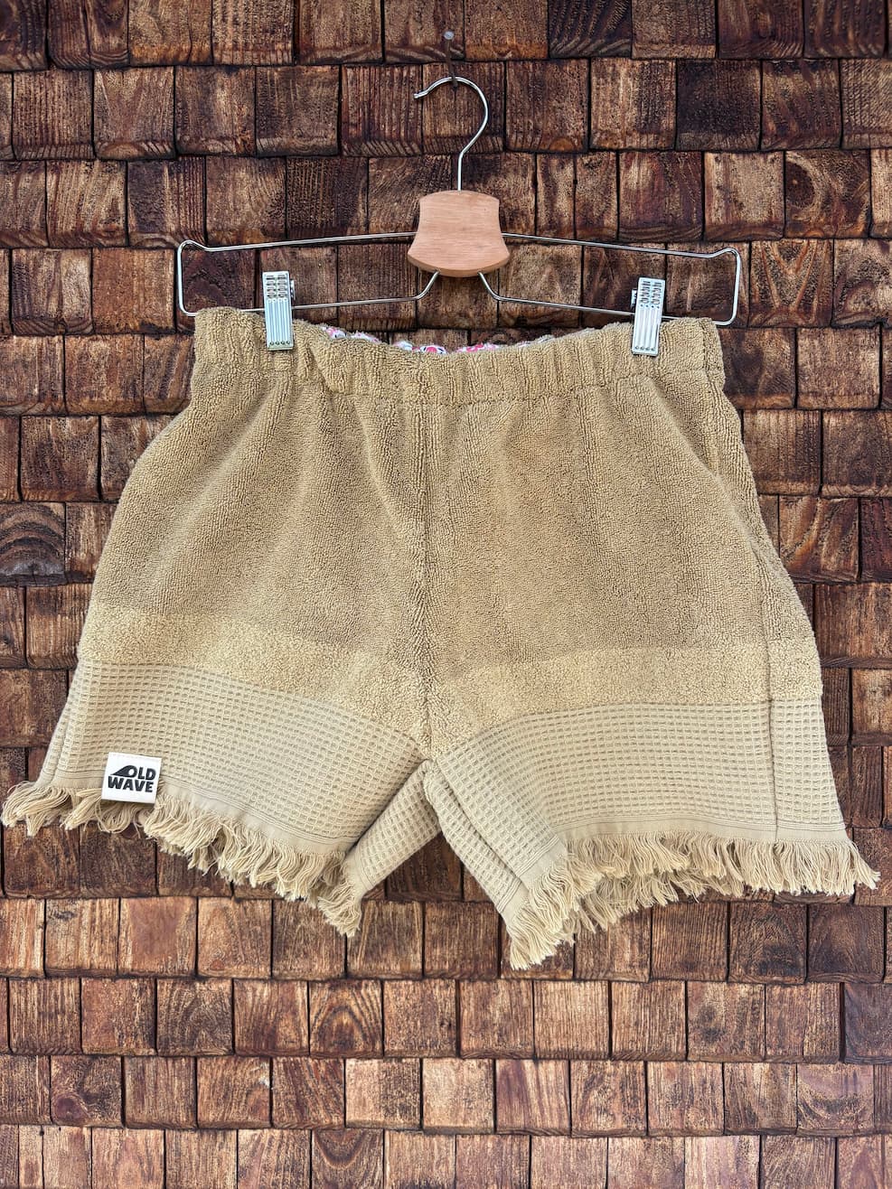Vorderansicht der Upcycling-Frotteeshorts „Sand Fringe ‘91“ von Old Wave auf einem Kleiderbügel vor einer rustikalen Holzwand. Die Shorts präsentieren ein schlichtes, einfarbiges Design in warmen Sand- und Beigetönen. Das linke Hosenbein ist am gefransten Saum mit dem Old Wave-Logo versehen. Der weiche Vintage-Stoff ist horizontal zweigeteilt in einen feinen oberen Bereich und eine strukturiertere untere Hälfte.