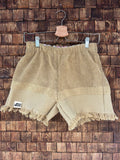 Vorderansicht der Upcycling-Frotteeshorts „Sand Fringe ‘91“ von Old Wave auf einem Kleiderbügel vor einer rustikalen Holzwand. Die Shorts präsentieren ein schlichtes, einfarbiges Design in warmen Sand- und Beigetönen. Das linke Hosenbein ist am gefransten Saum mit dem Old Wave-Logo versehen. Der weiche Vintage-Stoff ist horizontal zweigeteilt in einen feinen oberen Bereich und eine strukturiertere untere Hälfte.