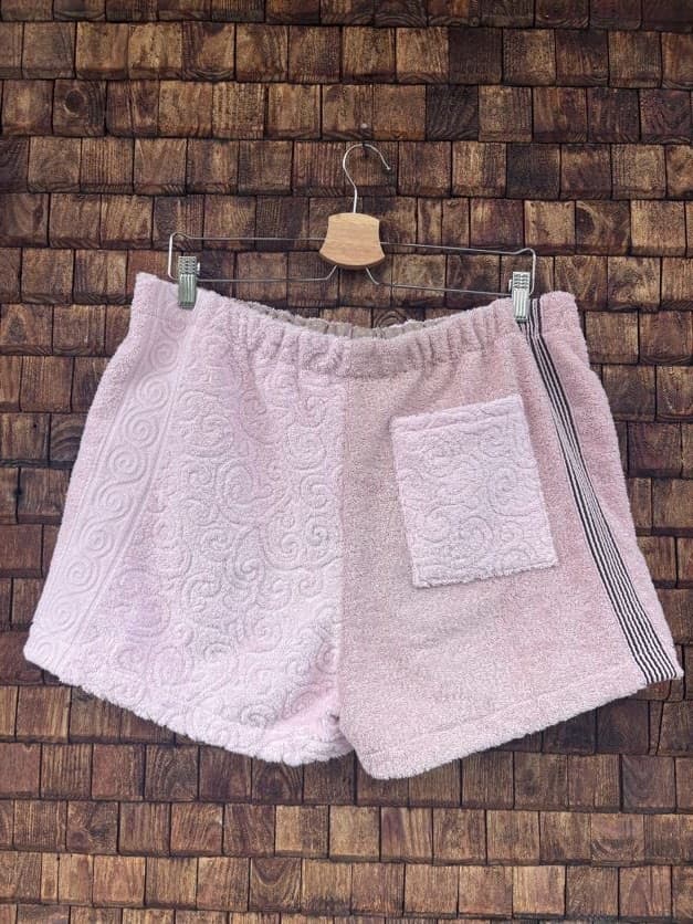 Rückansicht der Upcycling-Frotteeshorts „Sand Relief ‘103“ von Old Wave auf einem Kleiderbügel vor einer Holzwand. Die Rückseite zeigt die Aufteilung in eine rosa linke Gesäßhälfte und eine sand-beige rechte Hälfte mit dem grauen Seitenstreifen. Auf der rechten Seite ist zusätzlich eine quadratische, weiße Gesäßtasche aus Frottee aufgenäht.