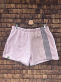 Vorderansicht der Upcycling-Frotteeshorts „Sand Relief ‘103“ von Old Wave auf einem Kleiderbügel vor einer rustikalen Holzwand. Das minimalistische Patchwork-Design kombiniert ein linkes Hosenbein in zartem Pastellrosa mit dem eingenähten Old Wave-Logo am Saum und ein rechtes Hosenbein in hellem Sand-Beige, das durch einen vertikalen grauen Streifen am äußeren Rand akzentuiert wird.