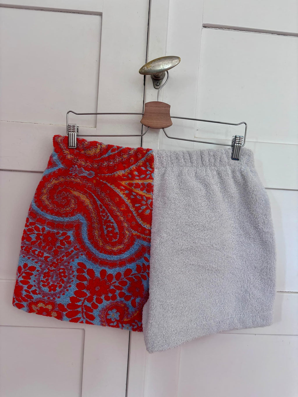 Rückansicht der Frottee-Patchwork-Shorts „Sardinia Summer 72“ von Old Wave. Zu sehen ist die Kombination aus zwei unterschiedlichen Vintage-Handtuchstoffen: Ein Hosenbein führt das lebendige rote Paisley-Muster fort, während das andere Hosenbein ein dezentes, weiß-hellblau feingestreiftes Muster zeigt.