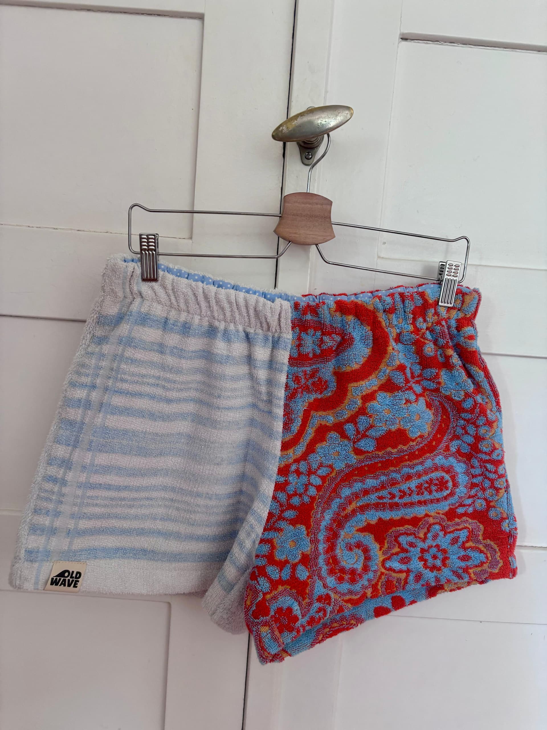 Upcycling-Shorts „Sardinia Summer 72“ von Old Wave an einem Kleiderbügel vor einer weißen Holztür. Das auffällige Patchwork-Design besteht aus einem komplett hellgrauen Hosenbein auf der einen Seite und einem kontrastierenden Hosenbein auf der anderen Seite aus rotem Frottee-Handtuchstoff mit einem großflächigen, geschwungenen Paisley-Muster in Hellblau und Orange.