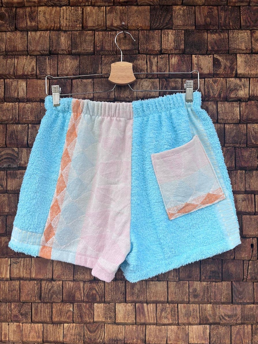 Rückansicht der Upcycling-Frotteeshorts „Sea Diamond ‘96“ von Old Wave auf einem Kleiderbügel vor einer Holzwand. Das asymmetrische Design zeigt auf der linken Gesäßhälfte einen dreifarbigen Patchwork-Mix aus Türkis, Weiß und Rosa mit einem vertikalen orangefarbenen Webstreifen, während die rechte, türkisfarbene Seite mit einer aufgesetzten Tasche im passenden hellrosa-weißen Muster gestaltet ist.