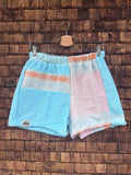 Vorderansicht der Upcycling-Frotteeshorts „Sea Diamond ‘96“ von Old Wave auf einem Kleiderbügel vor einer rustikalen Holzwand. Das maritime Pastell-Design kombiniert ein linkes Hosenbein aus hellem, unifarbenem Türkisblau mit dem typischen Old Wave-Logo am Saum sowie ein rechtes Hosenbein aus einem zartrosa-weißen Patchwork-Mix, der am Bund durch feine orangefarbene Musterakzente ergänzt wird.