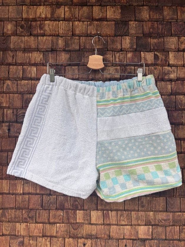 Rückansicht der Upcycling-Frotteeshorts „Sea Grid ‘105“ von Old Wave auf einem Kleiderbügel vor einer Holzwand. Das Patchwork-Muster spiegelt sich auf der Rückseite: Die linke Gesäßhälfte ist komplett in weißem Frotteestoff gehalten, während die rechte Hälfte das maritime mintgrün-türkise Muster mit horizontalen Streifen- und Rautenstrukturen zeigt.