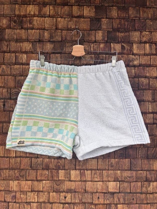 Vorderansicht der Upcycling-Frotteeshorts „Sea Grid ‘105“ von Old Wave auf einem Kleiderbügel vor einer rustikalen Holzwand. Das asymmetrische Design kombiniert ein linkes Hosenbein mit einem gemusterten Retro-Gitterstruktur-Stoff in Mintgrün und Türkis sowie dem eingenähten Old Wave-Logo am Saum, während das rechte Hosenbein schlicht aus unifarbenem, weißem Vintage-Frottee besteht.
