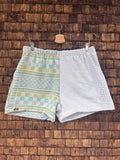 Vorderansicht der Upcycling-Frotteeshorts „Sea Grid ‘105“ von Old Wave auf einem Kleiderbügel vor einer rustikalen Holzwand. Das asymmetrische Design kombiniert ein linkes Hosenbein mit einem gemusterten Retro-Gitterstruktur-Stoff in Mintgrün und Türkis sowie dem eingenähten Old Wave-Logo am Saum, während das rechte Hosenbein schlicht aus unifarbenem, weißem Vintage-Frottee besteht.