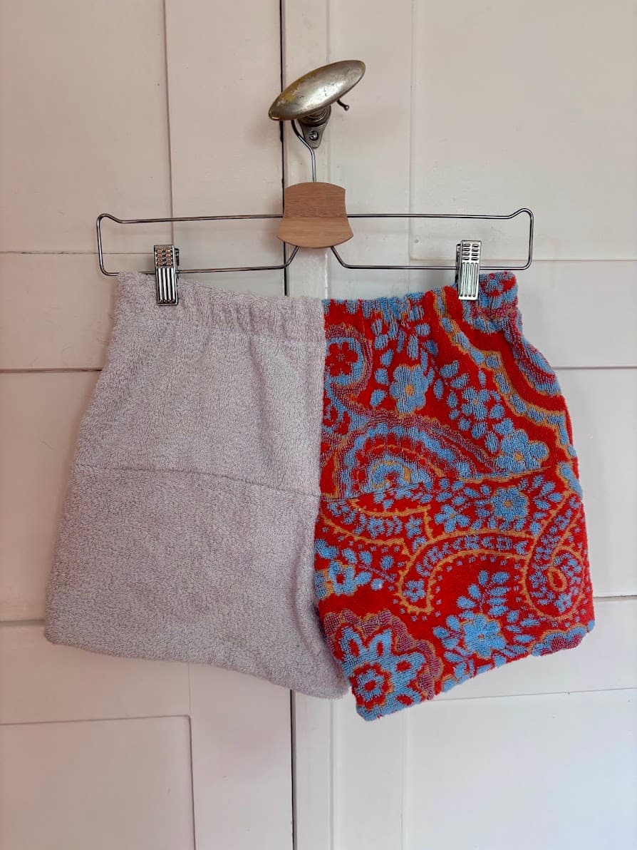Rückansicht der Frottee-Patchwork-Shorts „Sicilian Drift 64“ von Old Wave an einem Kleiderbügel. Diese Ansicht zeigt ein komplett hellgraues Hosenbein auf der linken Seite, kombiniert mit einem kontrastierenden rechten Hosenbein aus rotem Vintage-Handtuchstoff mit einem großflächigen, hellblauen Retro-Muster.