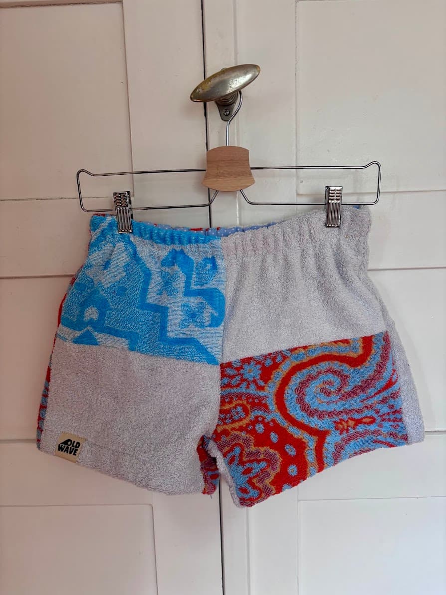 Upcycling-Shorts „Sicilian Drift 64“ von Old Wave auf einem Kleiderbügel vor einer weißen Holzwand. Die Vorderseite zeigt ein dreigeteiltes Patchwork-Design aus Frottee-Handtüchern: Ein hellgraues Hosenbein trifft auf ein geteiltes Hosenbein mit azurblauem Ornamentmuster im oberen Bereich und einem roten Paisley-Muster unten rechts.