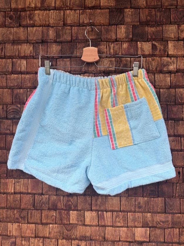 Rückansicht der Upcycling-Frotteeshorts „Sunfade Candy ‘97“ von Old Wave auf einem Kleiderbügel vor einer Holzwand. Die Rückseite ist vorwiegend aus hellem, pastellblauem Frotteestoff gefertigt, wobei die rechte Gesäßhälfte einen farbigen Akzent durch ein gestreiftes Patchwork-Element in Gelb und Pink mit einer aufgesetzten Tasche setzt.
