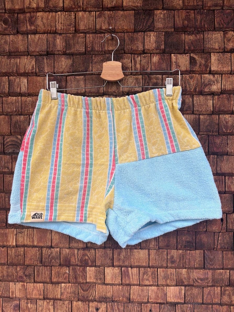 Vorderansicht der Upcycling-Frotteeshorts „Sunfade Candy ‘97“ von Old Wave auf einem Kleiderbügel vor einer rustikalen Holzwand. Das sommerliche Design kombiniert ein linkes Hosenbein mit feinen Längsstreifen in Gelb, Pink und Hellblau sowie dem Old Wave-Logo am Saum, während das rechte Hosenbein im unteren Bereich aus unifarbenem, hellblauem Vintage-Frottee besteht.