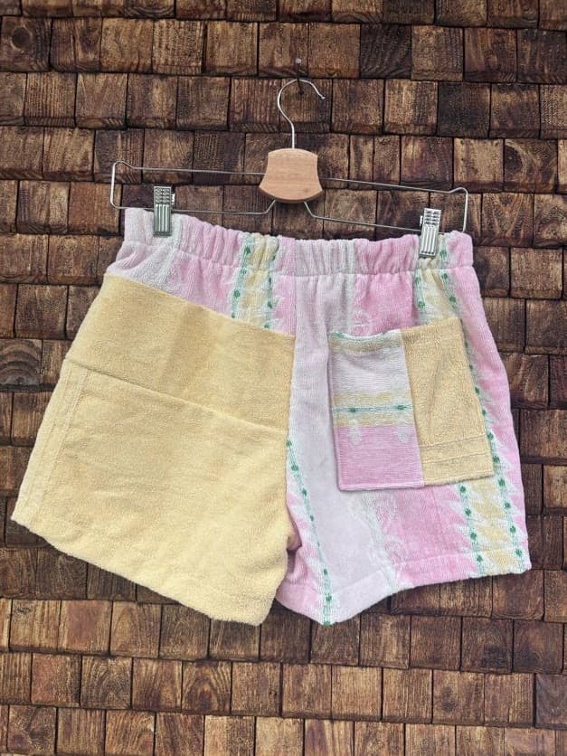 Rückansicht der Upcycling-Frotteeshorts „Sunfade Pastel ‘101“ von Old Wave auf einem Kleiderbügel vor einer Holzwand. Die Rückseite spiegelt das asymmetrische Patchwork-Design wider: Die linke Gesäßhälfte besteht aus dem unifarbenen, gelben Frotteestoff, während die rechte Hälfte das hellrosa-gestreifte Retromuster zeigt und mit einer aufgesetzten Tasche aus einem gelb-rosa-grünen Patchwork-Mix versehen ist.