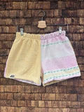 Vorderansicht der Upcycling-Frotteeshorts „Sunfade Pastel ‘101“ von Old Wave auf einem Kleiderbügel vor einer rustikalen Holzwand. Das zweigeteilte Pastell-Design zeigt ein linkes Hosenbein aus unifarbenem, sonnengelbem Vintage-Frottee mit dem eingenähten Old Wave-Logo am Saum, während das rechte Hosenbein aus einem hellen Stoff mit zarten, horizontalen Streifenstrukturen in Rosa, Mintgrün und Weiß gefertigt ist.
