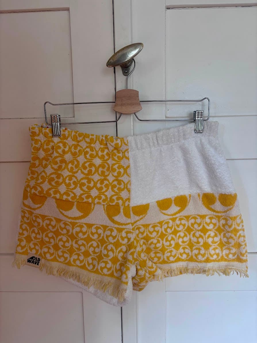 Upcycling-Shorts „Sunny Amalfi 70“ von Old Wave auf einem Kleiderbügel vor einer weißen Holzwand. Das Design aus Frottee-Handtüchern kombiniert eine rein weiße Partie oben rechts mit lebendigen, sonnengelben Mustern auf der linken Seite und entlang des unteren Saums, bestehend aus floralen Retro-Kacheln und bogenförmigen Ornamenten.