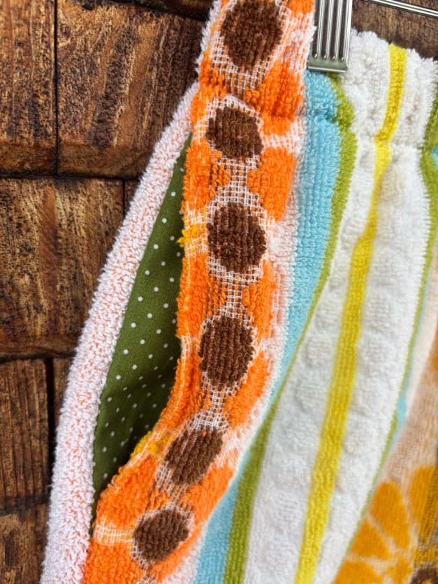Eine Nahaufnahme der Seitennaht und des Patchwork-Designs der „Sunny Daisy ‘73“ Frotteeshorts von Old Wave. Das Detail zeigt die kunstvolle Verbindung verschiedener Vintage-Frottee-Stoffe: Eine senkrechte Borte aus braunen Retro-Kreisen auf Orange wird flankiert von einem grün gepunkteten Stoff links mit rosa Fransen-Finish und einem türkisfarbenen Streifen rechts, neben weißem Frottee mit Waffelstruktur.