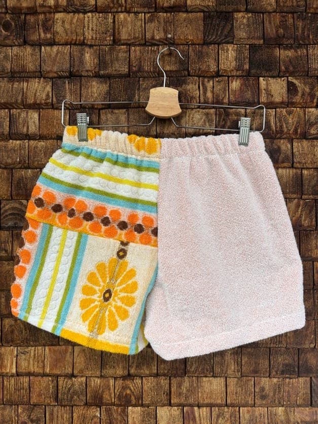 Rückansicht der Upcycling-Frotteeshorts „Sunny Daisy ‘73“ von Old Wave auf einem Kleiderbügel vor einer Holzwand. Die Rückseite besticht durch ein asymmetrisches Color-Blocking: Die linke Gesäßhälfte besteht komplett aus kuscheligem, unifarbenem Frotteestoff in Zartrosa, während die rechte Hälfte das lebendige Vintage-Muster aus gelben Blumen, türkisfarbenen Streifen und orangenen Retro-Kreisen fortführt.