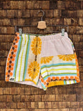 Vorderansicht der Upcycling-Frotteeshorts „Sunny Daisy ‘73“ von Old Wave auf einem Kleiderbügel vor einer rustikalen Holzwand. Das farbenfrohe 70er-Jahre-Retro-Design zeigt ein rechtes Hosenbein mit einer großen gelben Blume (Daisy) auf beigem Grund, umrahmt von bunten Streifen und Kreismustern in Orange und Türkis samt dem Logo-Patch. Das linke Hosenbein kombiniert im oberen Bereich zartrosa Frottee mit dem floralen Muster im unteren Teil.