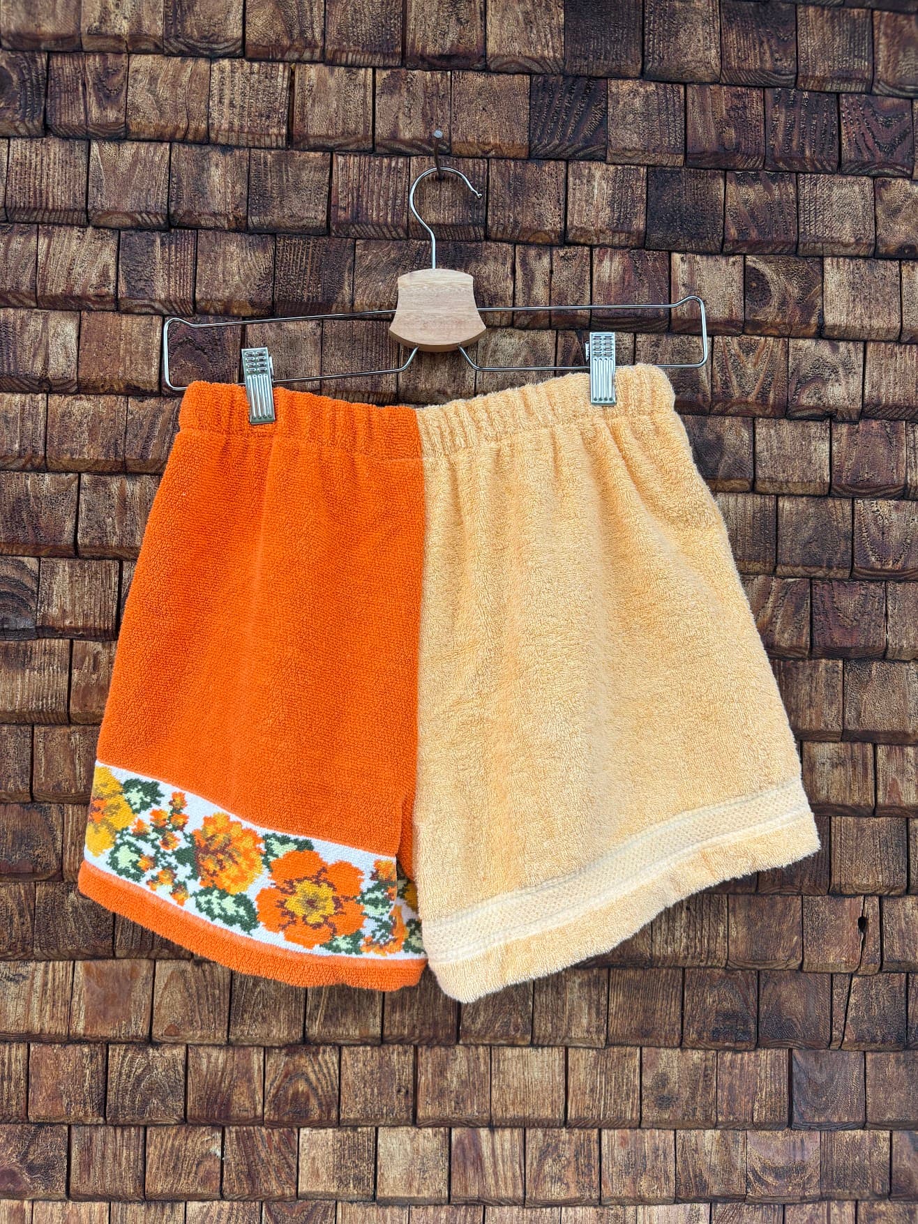 Rückansicht der Upcycling-Frotteeshorts „Sunshine Border ‘70“ von Old Wave auf einem Kleiderbügel vor einer Holzwand. Die Rückseite führt das asymmetrische Farbkonzept fort: Die linke Gesäßhälfte besteht aus unifarbenem, lebendigem Orange und zeigt am unteren Saum die durchlaufende, florale 70er-Jahre-Musterborte. Die rechte Gesäßhälfte ist minimalistisch aus weichem, hellgelbem Frotteestoff gefertigt.