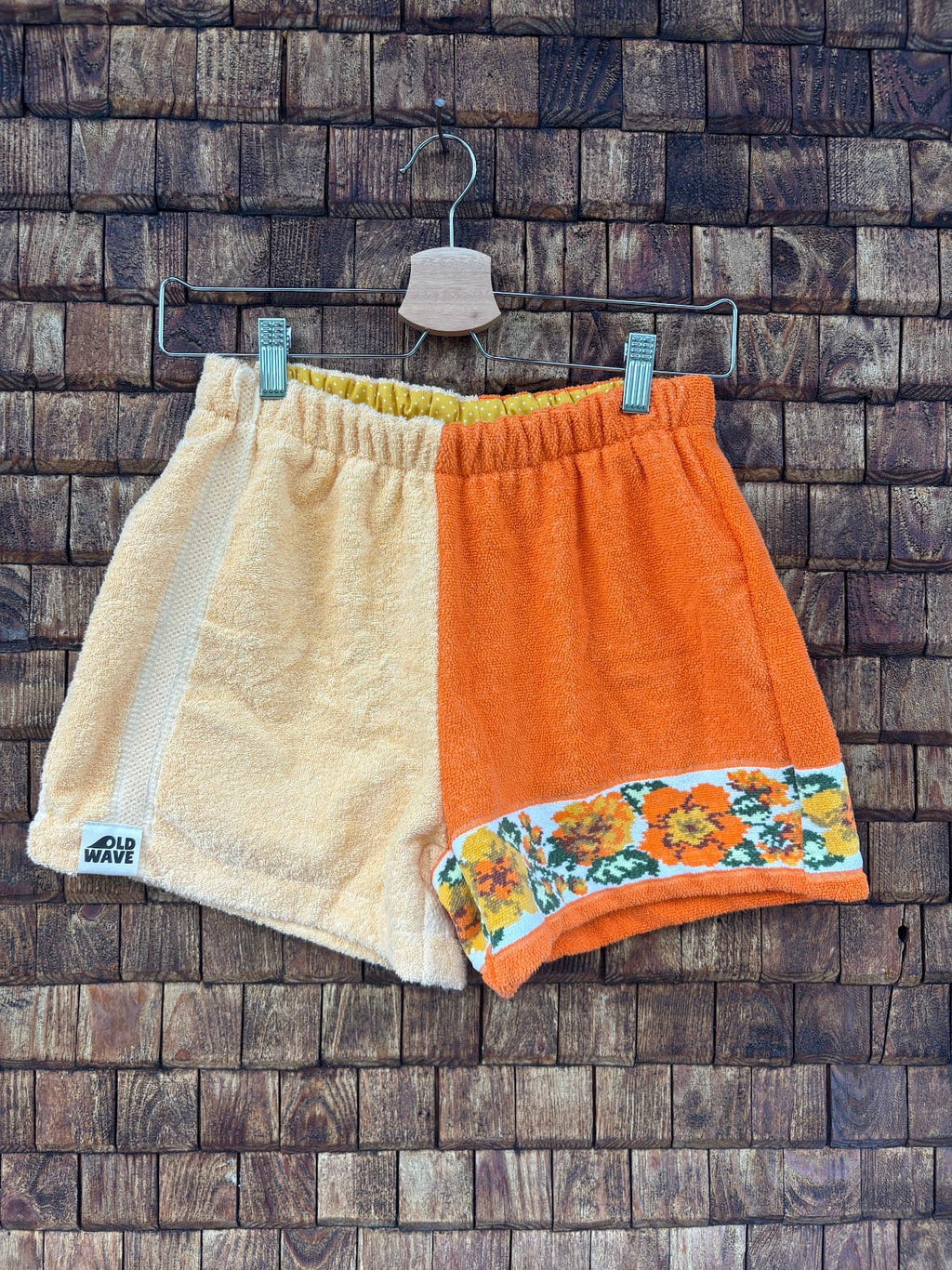 Vorderansicht der Upcycling-Frotteeshorts „Sunshine Border ‘70“ von Old Wave auf einem Kleiderbügel vor einer rustikalen Holzwand. Das zweigeteilte Retro-Design kombiniert ein rechtes Hosenbein aus hellgelbem Frottee samt Logo-Patch am Saum mit einem linken Hosenbein aus kräftigem Orange. Am unteren Saum der orangen Seite setzt eine nostalgische Musterborte mit gelb-orangefarbenen Retro-Blumen einen sommerlichen Akzent.