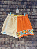 Vorderansicht der Upcycling-Frotteeshorts „Sunshine Border ‘70“ von Old Wave auf einem Kleiderbügel vor einer rustikalen Holzwand. Das zweigeteilte Retro-Design kombiniert ein rechtes Hosenbein aus hellgelbem Frottee samt Logo-Patch am Saum mit einem linken Hosenbein aus kräftigem Orange. Am unteren Saum der orangen Seite setzt eine nostalgische Musterborte mit gelb-orangefarbenen Retro-Blumen einen sommerlichen Akzent.