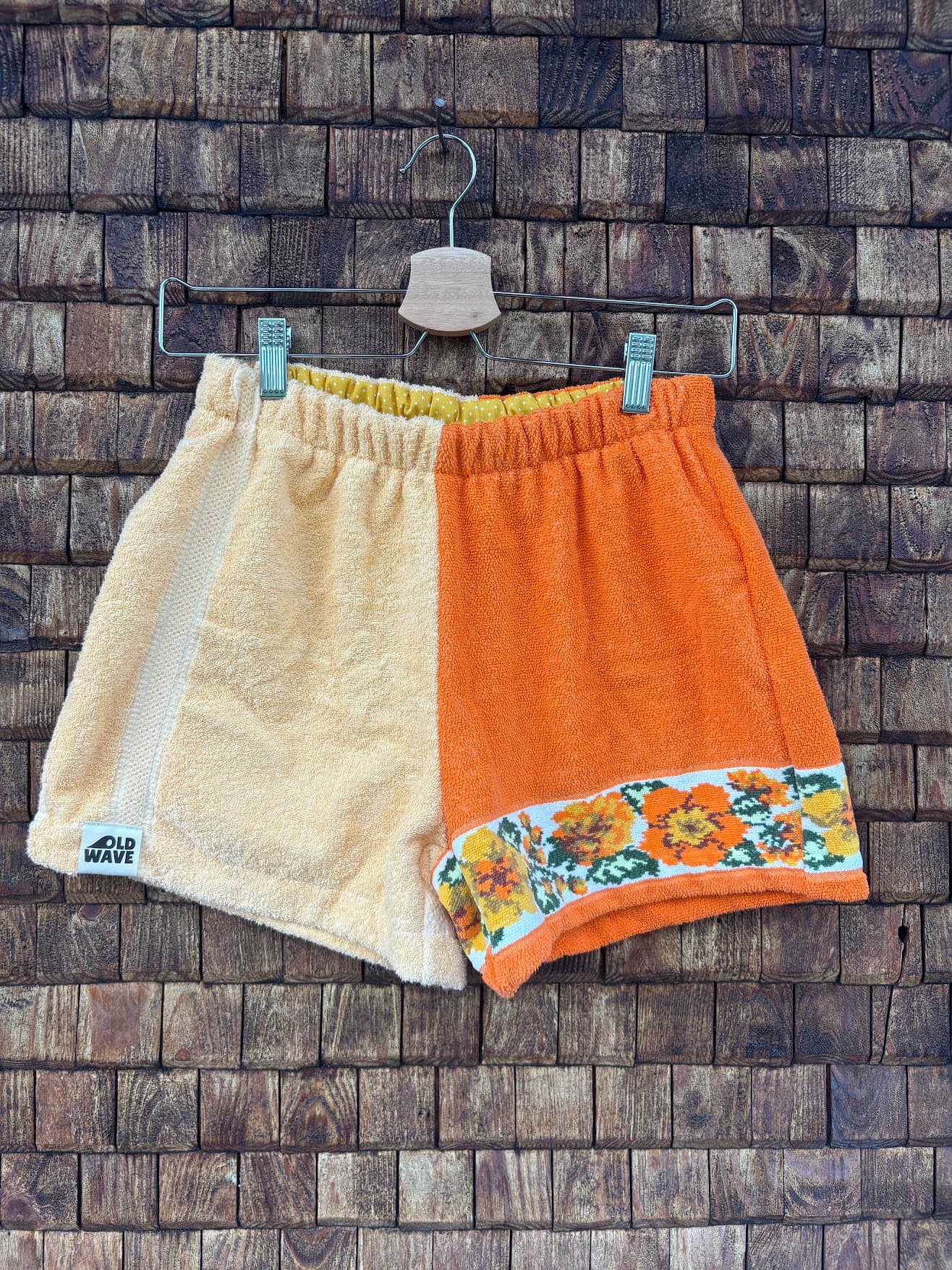 Vorderansicht der Upcycling-Frotteeshorts „Sunshine Border ‘70“ von Old Wave auf einem Kleiderbügel vor einer rustikalen Holzwand. Das zweigeteilte Retro-Design kombiniert ein rechtes Hosenbein aus hellgelbem Frottee samt Logo-Patch am Saum mit einem linken Hosenbein aus kräftigem Orange. Am unteren Saum der orangen Seite setzt eine nostalgische Musterborte mit gelb-orangefarbenen Retro-Blumen einen sommerlichen Akzent.
