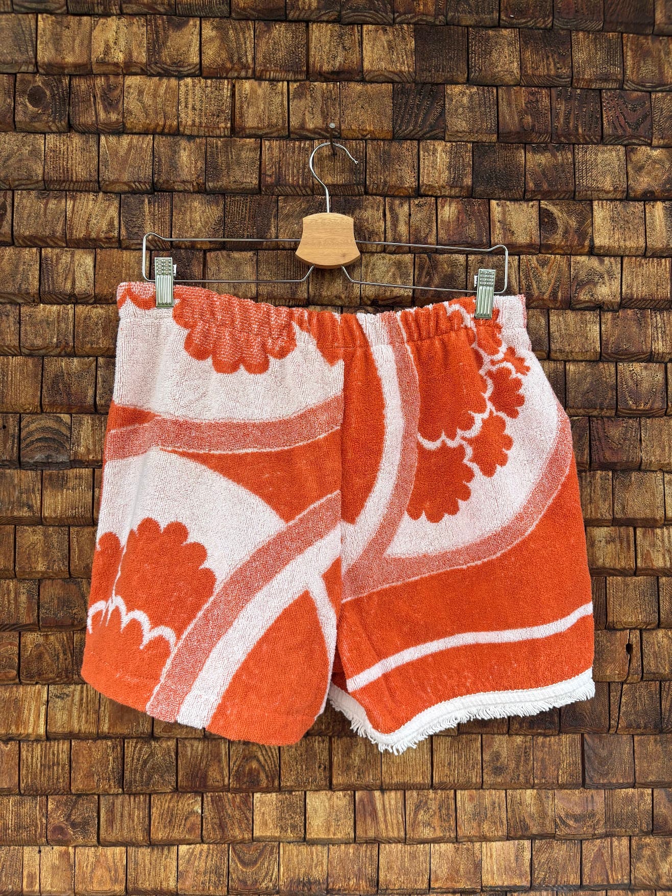 Rückansicht der Upcycling-Frotteeshorts „The Clean Cut“ (#3/5) von Old Wave auf einem Kleiderbügel vor einer Holzwand. Die Rückseite setzt das farbenfrohe All-over-Vintage-Muster nahtlos fort: Auf beiden Gesäßhälften verlaufen großformatige, orange-weiße Blumenornamente und geschwungene Linien mit einem leicht gefransten, weißen Saumabschluss.