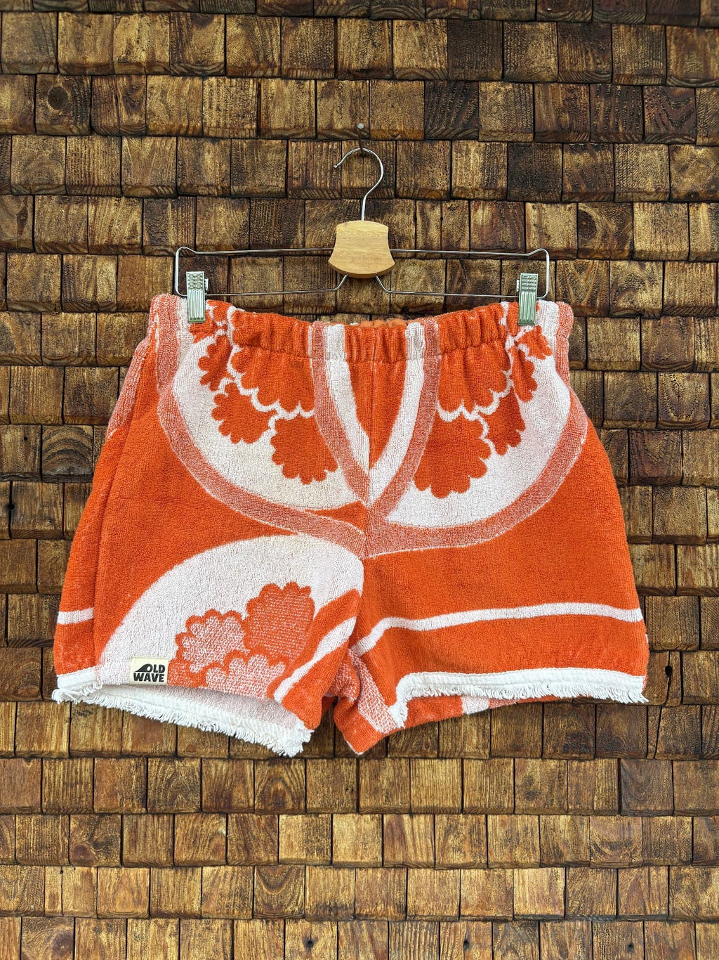 Vorderansicht der Upcycling-Frotteeshorts „The Clean Cut“ (#3/5) von Old Wave auf einem Kleiderbügel vor einer rustikalen Holzwand. Das Modell verzichtet auf Waffelpique-Einsätze und präsentiert ein klares, symmetrischer wirkendes Allover-Design aus kräftig orange-weißem Vintage-Frottee. Die Front besticht durch geschwungene, großflächige Retro-Blumenmotive und trägt das dezente Old Wave Logo-Patch am Saum des rechten Hosenbeins.
