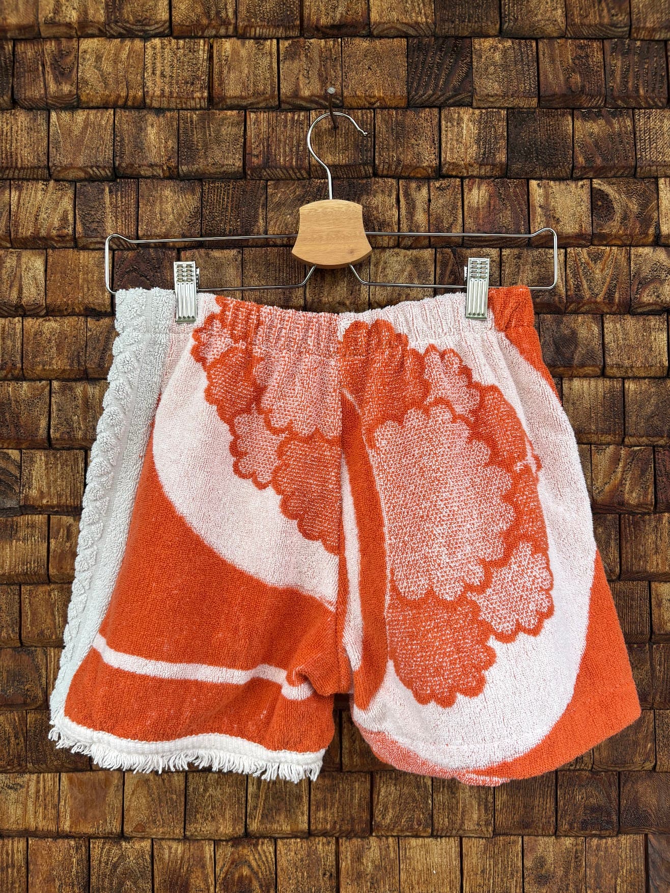 Rückansicht der Upcycling-Frotteeshorts „The Patchwork Twin“ von Old Wave auf einem Kleiderbügel vor einer Holzwand. Die Rückseite spiegelt das asymmetrische Design wider: Die linke Gesäßhälfte besteht aus dem strukturierten, weißen Waffelpique-Frottee, während die rechte Gesäßhälfte das lebendige, orange-weiße Retro-Blumenmuster aufgreift. Beide Hosenbeine haben einen gefransten Saum.