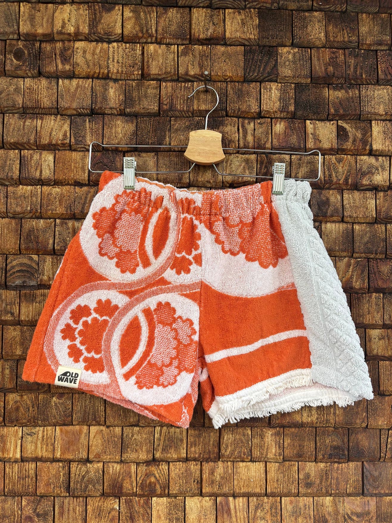 Vorderansicht der Upcycling-Frotteeshorts „The Patchwork Twin“ von Old Wave auf einem Kleiderbügel vor einer rustikalen Holzwand. Das asymmetrische Patchwork-Design kombiniert ein rechtes Hosenbein aus weißem Waffelpique-Frottee mit einem linken Hosenbein aus orangefarbenem Vintage-Frottee, das mit großen, weißen, grafischen Blumenmotiven verziert ist.