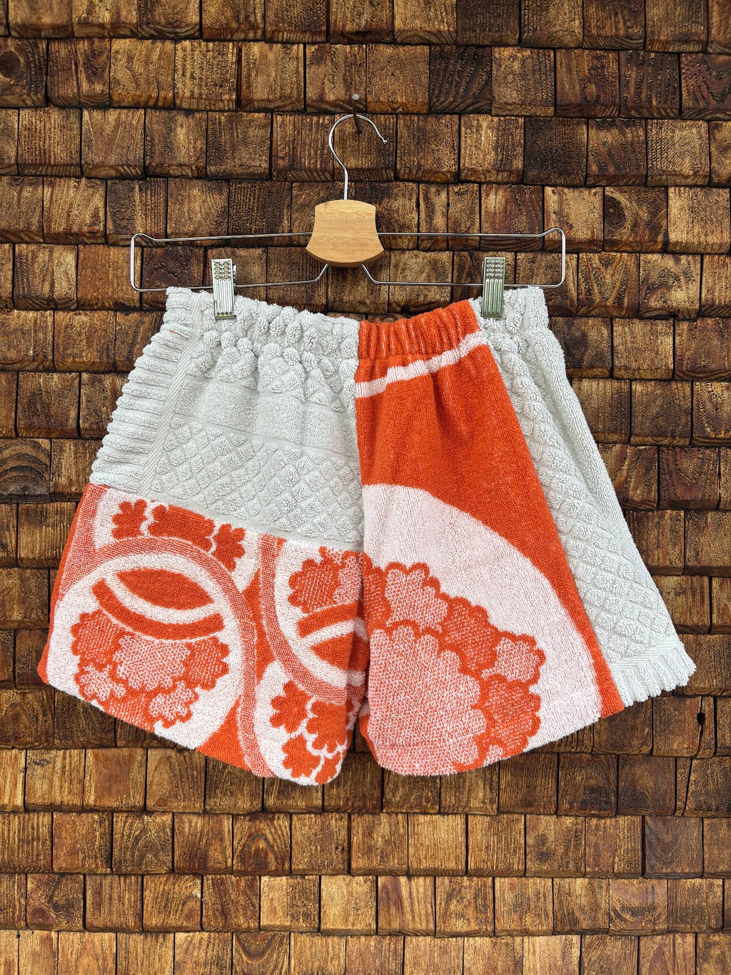 Rückansicht der Upcycling-Frotteeshorts „The Retro Sidepanel“ (#4/5) von Old Wave auf einem Kleiderbügel vor einer Holzwand. Das asymmetrische Patchwork-Design zeigt auf der linken Gesäßhälfte oben weißes Waffelpique-Frottee und unten ein orange-weißes Blütenmuster. Die rechte Gesäßhälfte wird von einem großen, geschwungenen floralen Retro-Motiv in Orange dominiert, das seitlich von einem weißen Sidepanel eingefasst wird.