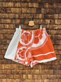 Vorderansicht der Upcycling-Frotteeshorts „The Retro Sidepanel“ (#4/5) von Old Wave auf einem Kleiderbügel vor einer rustikalen Holzwand. Das Design besticht durch ein dominantes Mittelteil aus orange-weißem Vintage-Frottee mit auffälligen, kreisförmigen Blumenmotiven samt Logo-Patch am Saum. Ein seitliches Panel aus strukturiertem, weißem Waffelpique-Frottee setzt am rechten Hosenbein einen sportlichen Kontrast.