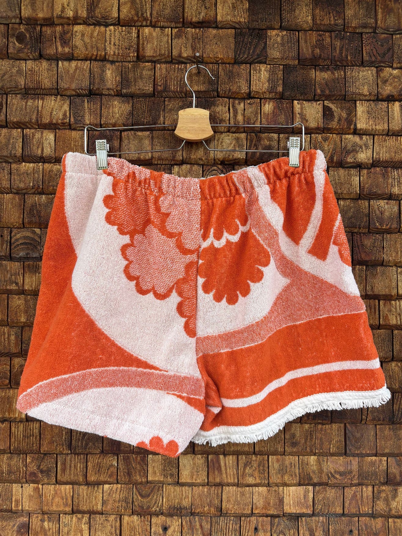 Rückansicht der Upcycling-Frotteeshorts „The Statement Piece“ (#2/5) von Old Wave auf einem Kleiderbügel vor einer Holzwand. Die Rückseite führt das ausdrucksstarke Vintage-Muster fort: Die linke Gesäßhälfte zeigt eine große, geschwungene Blumen-Grafik in sattem Orange auf weißem Frottee, während das rechte Hosenbein durch ein vertikales, florales Muster-Panel und einen orangefarbenen Grundton mit gefranstem Saum dominiert wird.