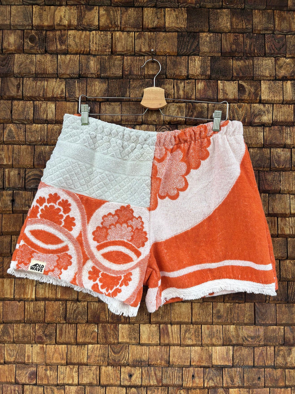 Vorderansicht der Upcycling-Frotteeshorts „The Statement Piece“ (#2/5) von Old Wave auf einem Kleiderbügel vor einer rustikalen Holzwand. Das kreative Patchwork-Design besticht durch ein rechtes Hosenbein, das im oberen Teil aus weißem Waffelpique-Frottee und unten aus einem orange-weißen Retro-Blumenmuster samt Logo-Patch am Saum besteht. Das linke Hosenbein zeigt ein großflächiges, florales Vintage-Motiv in kräftigem Orange auf weißem Grund.