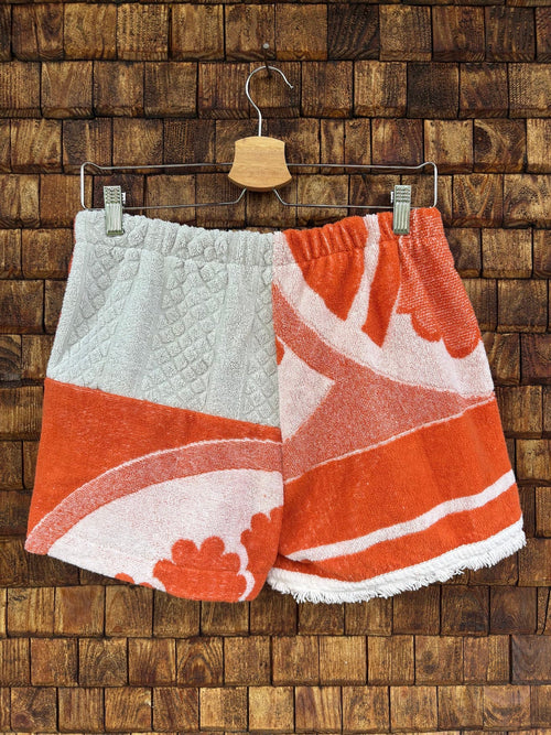 Rückansicht der Upcycling-Frotteeshorts „The Sunburst“ (#5/5) von Old Wave auf einem Kleiderbügel vor einer Holzwand. Das asymmetrische Patchwork-Design kombiniert auf der linken Gesäßhälfte ein oberes Feld aus strukturiertem, weißem Waffelpique-Frottee mit einem orange-weißen Blumenmuster unten. Die rechte Gesäßhälfte zeigt großflächige, geschwungene Retro-Motive in kräftigem Orange auf weißem Grund.