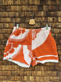 Vorderansicht der Upcycling-Frotteeshorts „The Sunburst“ (#5/5) von Old Wave auf einem Kleiderbügel vor einer rustikalen Holzwand. Das sonnige Vintage-Design besticht durch leuchtend orange-weiße, geschwungene Retro-Blumen- und Strahlenmotive über beide Hosenbeine sowie das Old Wave Logo-Patch am Saum des rechten Hosenbeins.