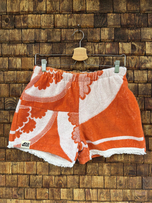 Vorderansicht der Upcycling-Frotteeshorts „The Sunburst“ (#5/5) von Old Wave auf einem Kleiderbügel vor einer rustikalen Holzwand. Das sonnige Vintage-Design besticht durch leuchtend orange-weiße, geschwungene Retro-Blumen- und Strahlenmotive über beide Hosenbeine sowie das Old Wave Logo-Patch am Saum des rechten Hosenbeins.