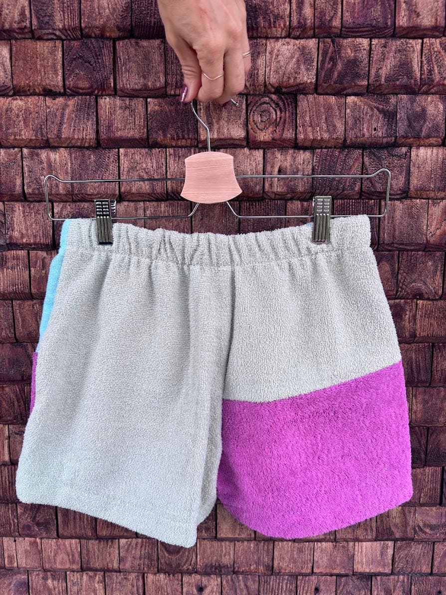 Rückansicht der Frottee-Patchwork-Shorts „Vanilla '66“ von Old Wave an einem Kleiderbügel. Diese Perspektive zeigt das asymmetrische Gesäßdesign aus upgecycelten Vintage-Handtüchern: Die Rückseite besteht überwiegend aus einem weichen, mintfarbenen Frotteestoff, wobei das rechte Hosenbein im unteren Drittel durch eine kontrastierende Stoffpartie in kräftigem Flieder abgeschlossen wird.