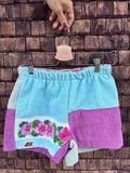 Upcycling-Shorts „Vanilla '66“ von Old Wave auf einem Kleiderbügel vor einer dunklen Holzwand. Das Patchwork-Design aus Frottee-Handtüchern kombiniert ein rechtes Hosenbein in kräftigem Flieder mit einem hellblauen linken Hosenbein, das im unteren Bereich eine geschwungene weiße Bordüre mit leuchtend rosa-pinken Blüten zeigt und an einen seitlichen mintfarbenen Streifen grenzt.