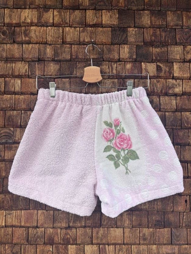 Rückansicht der Upcycling-Frotteeshorts „Vintage Bouquet ‘107“ von Old Wave auf einem Kleiderbügel vor einer Holzwand. Das romantische Design in Pastellrosa setzt das florale Thema fort: Die rechte Gesäßhälfte zeigt einen vertikalen, weißen Patchwork-Streifen, auf dem eine eingewebte Rose mit grünen Blättern abgebildet ist, während die linke Seite schlicht rosa gehalten ist.