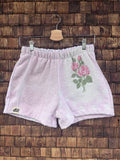Vorderansicht der Upcycling-Frotteeshorts „Vintage Bouquet ‘107“ von Old Wave auf einem Kleiderbügel vor einer rustikalen Holzwand. Die Shorts sind in zartem Pastellrosa gehalten, wobei die linke Seite am Saum das eingenähte Old Wave-Logo trägt. Auf dem rechten Hosenbein setzt ein weißer Patchwork-Einsatz mit einer eingewebten, rosa-grünen Vintage-Rosen-Stickerei einen floralen Akzent.