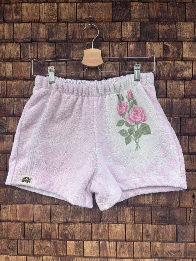 Vorderansicht der Upcycling-Frotteeshorts „Vintage Bouquet ‘107“ von Old Wave auf einem Kleiderbügel vor einer rustikalen Holzwand. Die Shorts sind in zartem Pastellrosa gehalten, wobei die linke Seite am Saum das eingenähte Old Wave-Logo trägt. Auf dem rechten Hosenbein setzt ein weißer Patchwork-Einsatz mit einer eingewebten, rosa-grünen Vintage-Rosen-Stickerei einen floralen Akzent.