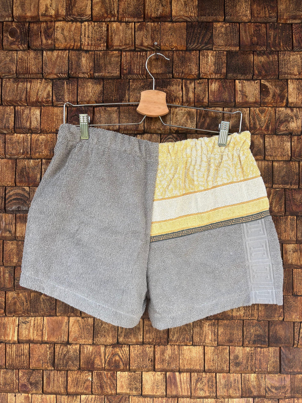 Rückansicht der Upcycling-Frotteeshorts „Vintage Grid & Grey ‘76“ von Old Wave auf einem Kleiderbügel vor einer Holzwand. Die Rückseite präsentiert ein asymmetrisches Color-Blocking-Design mit einer linken Hälfte aus schlichtem, meliertem grauem Frotteestoff. Die rechte Seite führt das gelbe Vintage-Gittermuster am Bund fort, das unten in einen grauen, gemusterten und einen unifarbenen grauen Frotteeabschnitt übergeht.