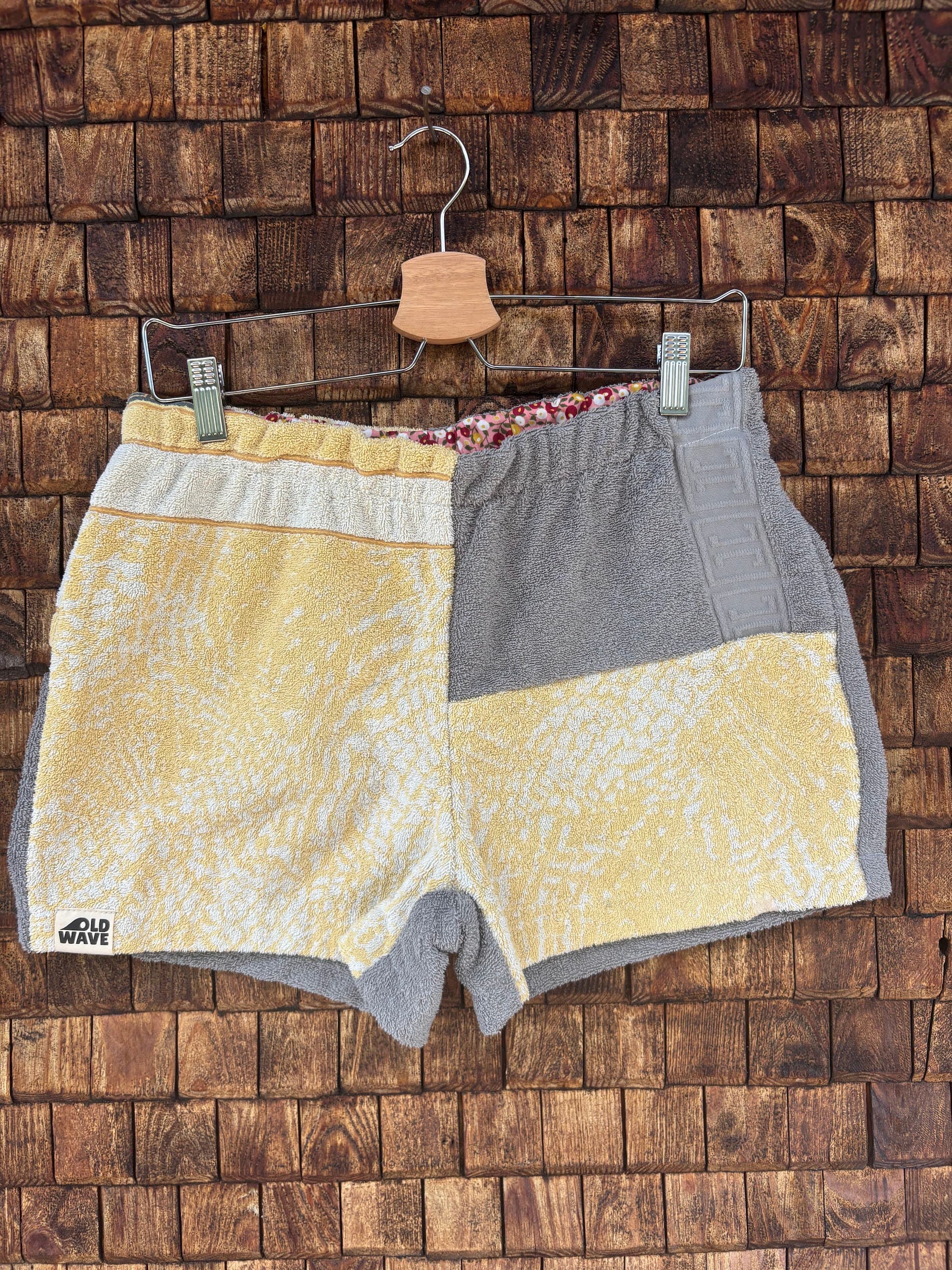 Vorderansicht der Upcycling-Frotteeshorts „Vintage Grid & Grey ‘76“ von Old Wave auf einem Kleiderbügel vor einer rustikalen Holzwand. Das Patchwork-Design besticht durch eine linke Seite aus sonnengelben Frottee mit einem dezenten, weißen Gittermuster (Grid) und dem Old Wave-Logo am Saum. Die rechte Gesäßseite kombiniert im oberen Bereich schlichtes, graues Frottee mit einem gelb-weiß-grauen Patchwork-Mix und einem gemusterten Bund.