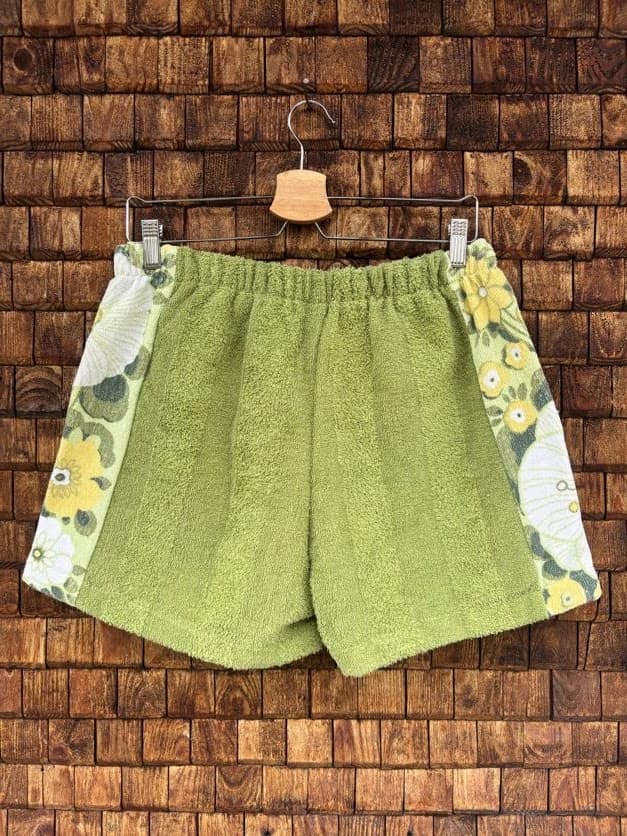 Rückansicht der Upcycling-Frotteeshorts „Wildflower Mix ‘79“ von Old Wave auf einem Kleiderbügel vor einer Holzwand. Die Rückseite besticht durch eine symmetrische Aufteilung, bei der das Hauptteil komplett aus unifarbenem, olivgrünem Frotteestoff gefertigt ist. An beiden Außenseiten setzen breite, vertikale Einsätze mit dem nostalgischen gelb-weißen Retro-Wildblumenmuster kontrastreiche Akzente.