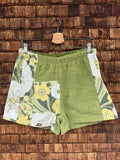 Vorderansicht der Upcycling-Frotteeshorts „Wildflower Mix ‘79“ von Old Wave auf einem Kleiderbügel vor einer rustikalen Holzwand. Das grüne Retro-Design kombiniert ein strukturiertes, olivgrünes Frottee-Zentrum mit verspielten Patchwork-Einsätzen. Das linke Hosenbein zeigt im unteren Bereich ein gelb-weißes Wildblumenmuster, während das rechte Hosenbein seitlich von einem diagonalen Musterstreifen in Grün- und Gelbtönen flankiert wird.