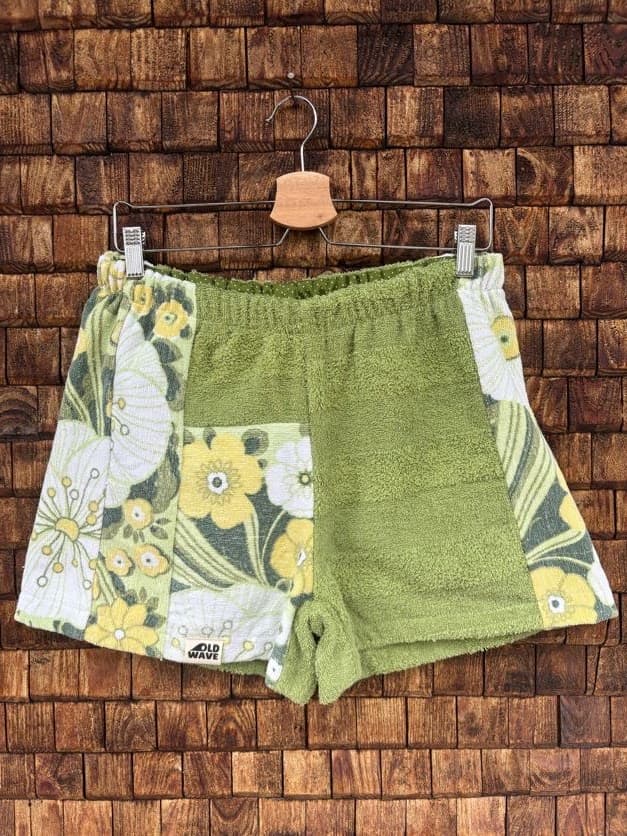 Vorderansicht der Upcycling-Frotteeshorts „Wildflower Mix ‘79“ von Old Wave auf einem Kleiderbügel vor einer rustikalen Holzwand. Das grüne Retro-Design kombiniert ein strukturiertes, olivgrünes Frottee-Zentrum mit verspielten Patchwork-Einsätzen. Das linke Hosenbein zeigt im unteren Bereich ein gelb-weißes Wildblumenmuster, während das rechte Hosenbein seitlich von einem diagonalen Musterstreifen in Grün- und Gelbtönen flankiert wird.